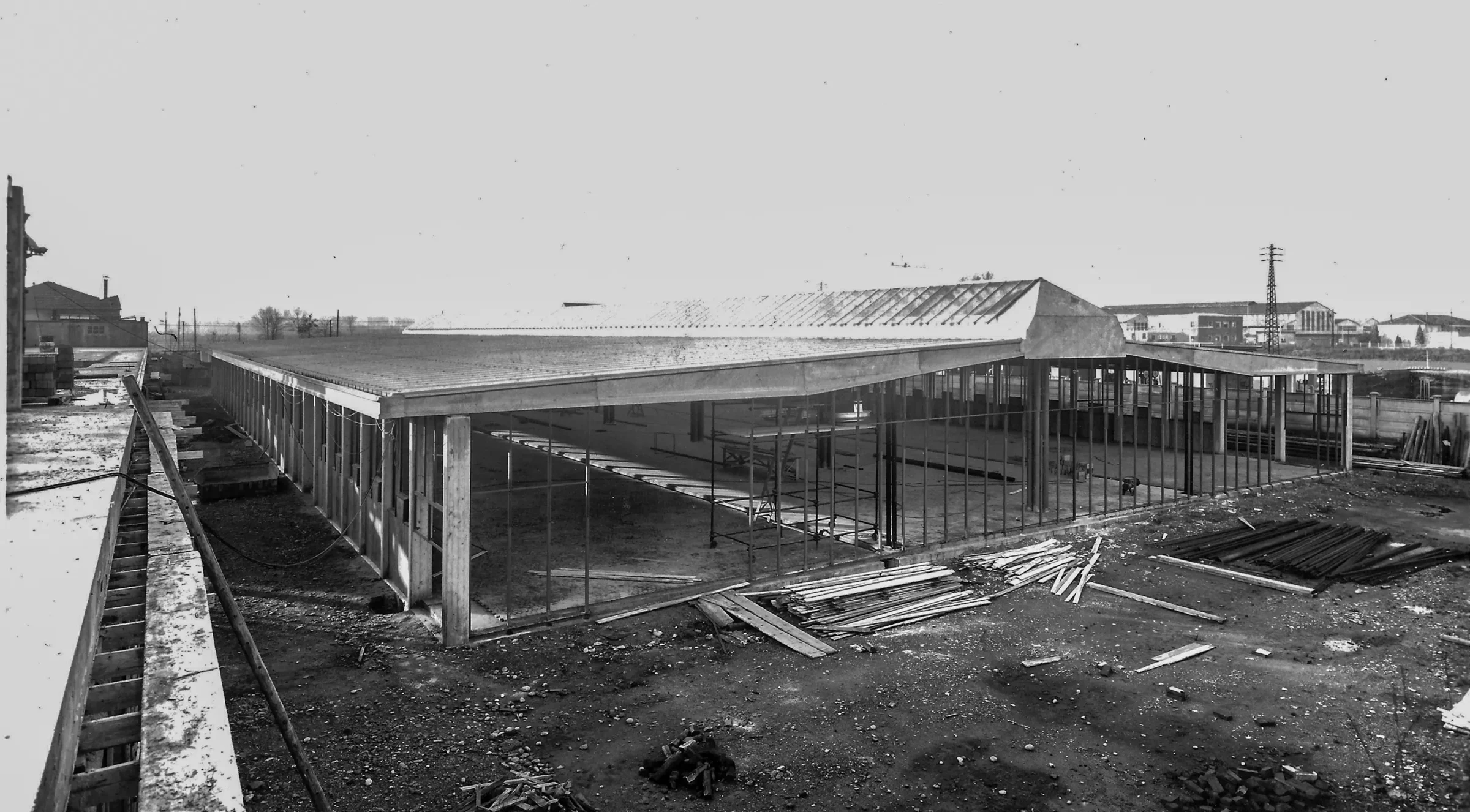 Black and white archival photo: the reinforced concrete structure of a one-story building, with the roof already installed and the construction site covered with panels and pipes.