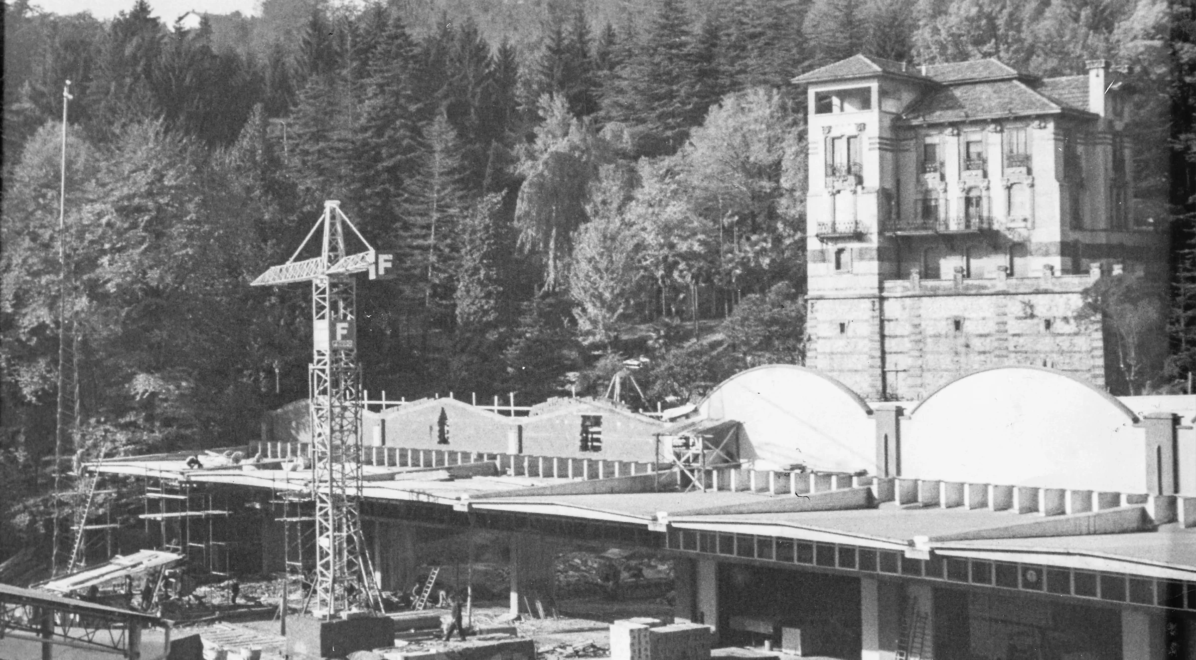 Black and white archive photo: a construction crane next to a building under construction at the foot of a wooded hill, with a historic multi-story villa in the background.